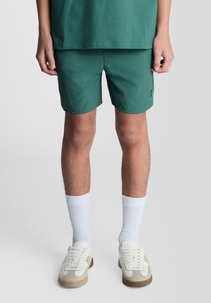 Man draagt groene korte broek, witte kousen en witte sneakers, staand tegen een lichtgrijze achtergrond.