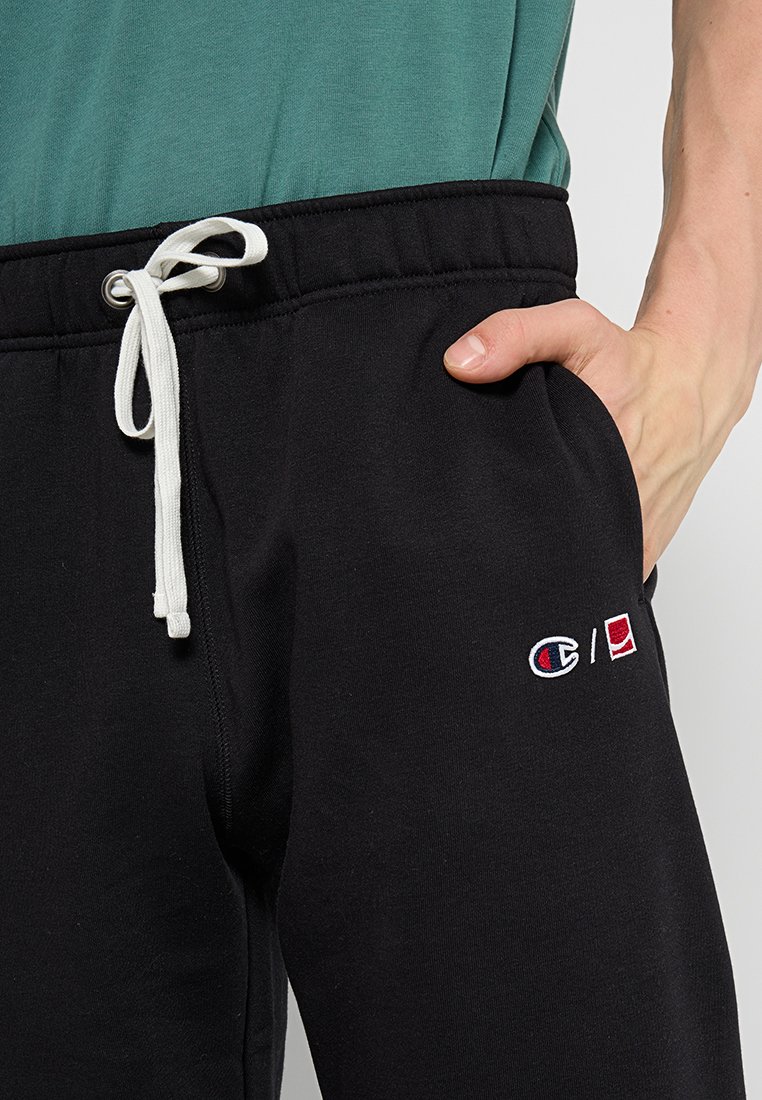 Pantaloni sportivi neri con un cordino bianco, caratterizzati da loghi ricamati in rosso e bianco sulla coscia sinistra. Tessuto di cotone morbido, vestibilità rilassata.