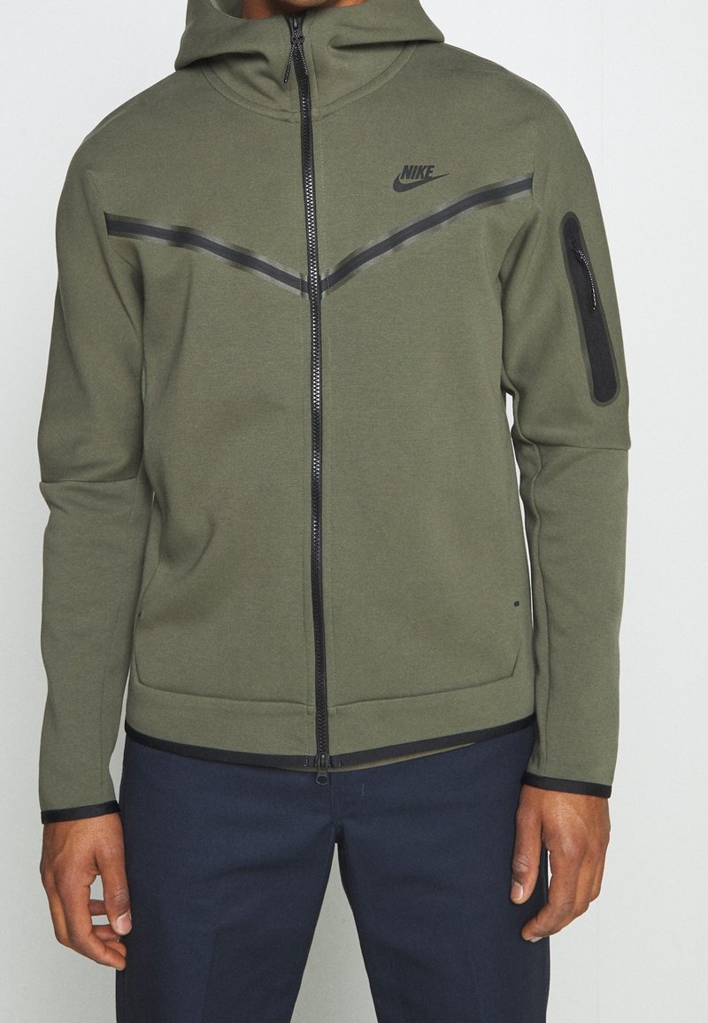 Person som bär en olivgrön Nike zip-up jacka med svarta chevronränder och en dragkedjeficka på vänster ärm, kombinerat med mörka byxor.