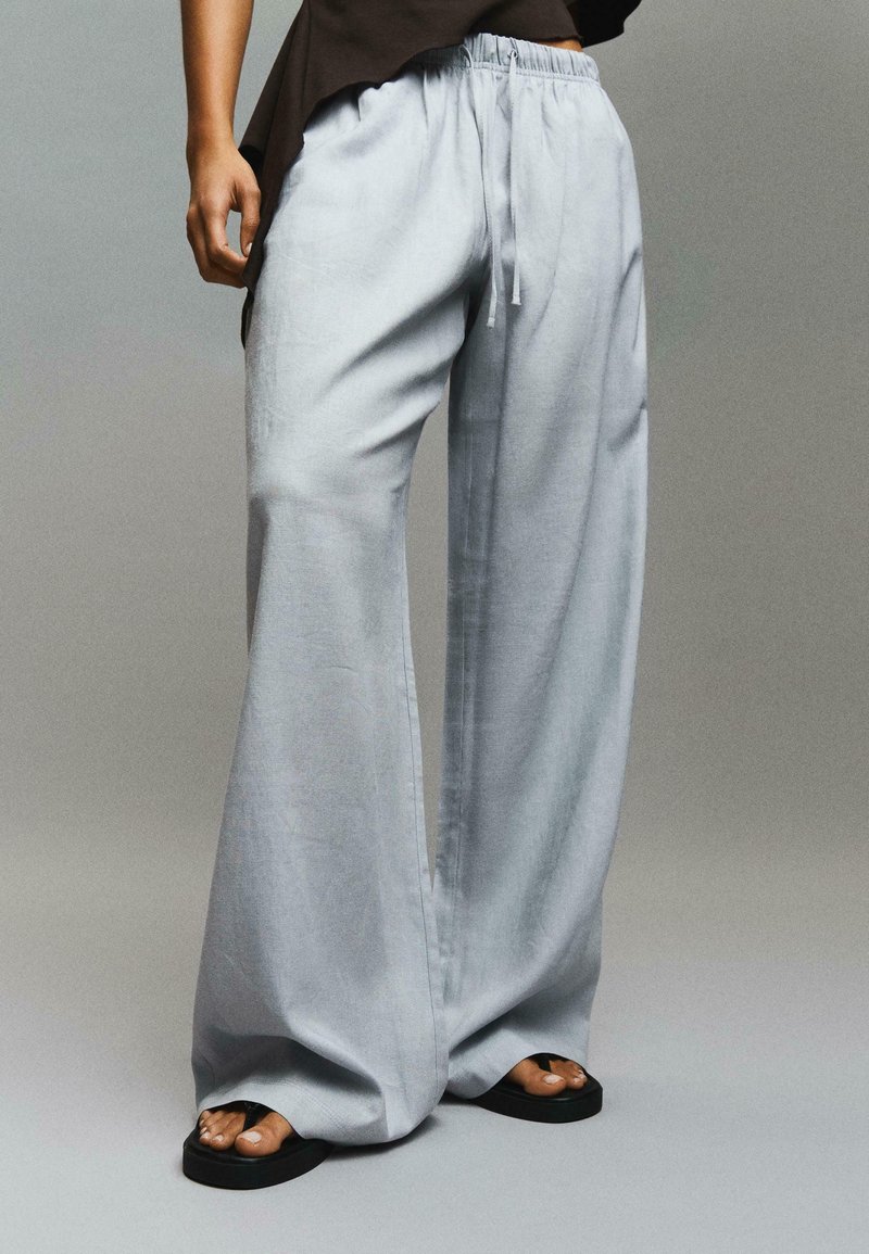 Personne portant un pantalon ample gris clair avec une taille à cordon et des sandales noires, debout devant un fond uni.
