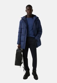 Mężczyzna w niebieskiej ocieplanej kurtce, niebieskim swetrze, ciemnych spodniach i czarnych butach, trzyma czarny plecak przy boku, stojący na białym tle.