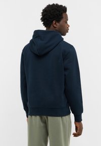 Mężczyzna w granatowej bluzie z kapturem i jasnoszarych spodniach, stojący i patrzący w prawo na tle jednolitego białego tła.