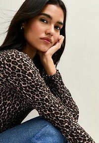 Top a maniche lunghe con stampa leopardata realizzato in tessuto morbido, caratterizzato da un design attillato. Il modello è seduto con una mano sul mento, indossando jeans in denim.