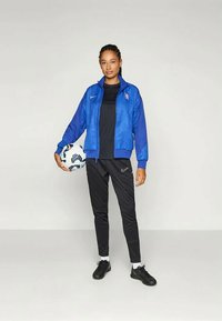 Modrá nike bunda s textúrovaným dizajnom, čierne tričko, čierne nike nohavice a čierne topánky. Drží biely futbalový loptu.