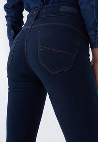 Un jean en denim bleu marine avec une coupe ajustée, des surpiqûres orange contrastantes, et des poches arrière ornées de coutures incurvées distinctives.