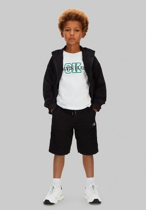 Niño con cabello rizado que lleva una chaqueta negra, camiseta blanca con el logo "Calvin Klein", pantalones cortos negros, calcetines blancos y zapatillas blancas, de pie con las manos en los bolsillos.