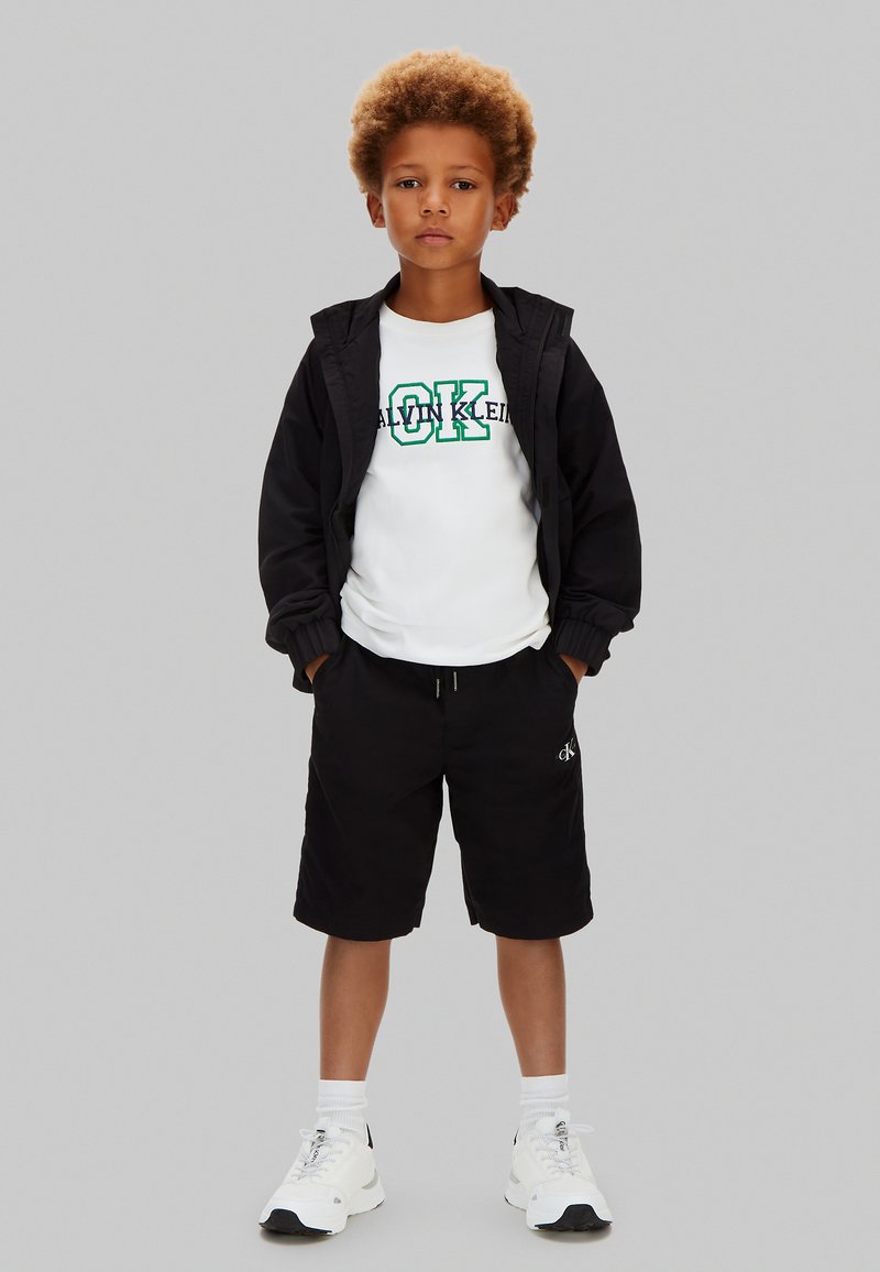 Niño con cabello rizado que lleva una chaqueta negra, camiseta blanca con el logo "Calvin Klein", pantalones cortos negros, calcetines blancos y zapatillas blancas, de pie con las manos en los bolsillos.
