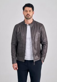 Mężczyzna w ciemnobrązowej skórzanej kurtce na białej koszuli i czarnych spodniach, stojący z jedną ręką w kieszeni na jednolitym tle.