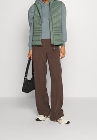 Grove groene bodywarmer over een grijze hoodie, bruine brede pijpen broek en witte sneakers. Zwarte schoudertas met metalen details.