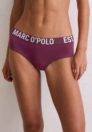 Frau in lila hochgeschnittenen Unterhosen mit dem "MARC O'POLO"-Logo auf dem elastischen Bund, stehend vor einer weißen strukturierten Wand.