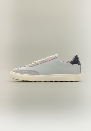 Zapatilla baja en gris claro con suela beige, cordones blanco roto, puntera de ante y lengüeta trasera oscura, mostrada de lado sobre un fondo liso.