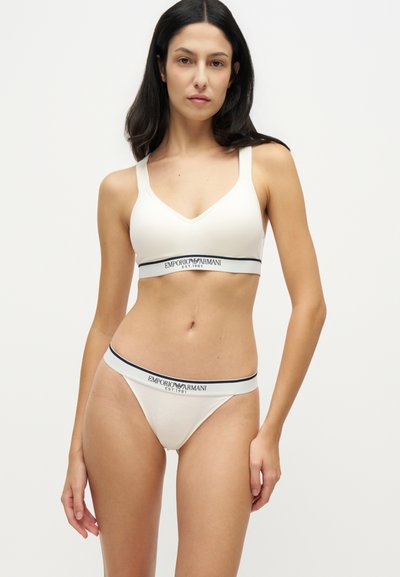 Γυναίκα που φοράει λευκό bralette Emporio Armani και αντίστοιχα εσώρουχα με λωρίδες λογότυπου, στέκεται μπροστά σε απλό ανοιχτό φόντο.