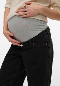 Personne enceinte portant un jean de maternité noir et un haut beige, les mains reposant doucement sur leur ventre.