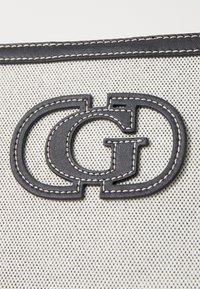 Sac en toile présentant un logo en cuir noir avec des coutures contrastantes sur un fond texturé de couleur claire. Lignes épurées et design moderne.