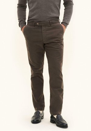 Man draagt bruine broek, grijze longsleeve, witte sokken en zwarte leren loafers, staat met handen in zakken.