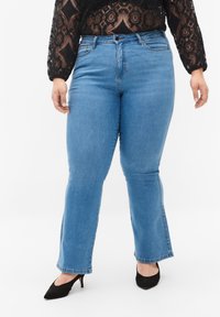 Jeans de campana azules con una cintura de altura media, bolsillos frontales y detalles cosidos en las costuras, combinados con una blusa de encaje negra y tacones negros.