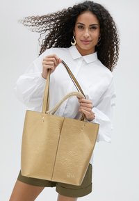 Zlatá textúrovaná taška s korálkovou rukoväťou, vertikálnymi švami a metalickým leskom. Skombinovaná s bielou košeľou s gombíkmi a olivovými šortkami.