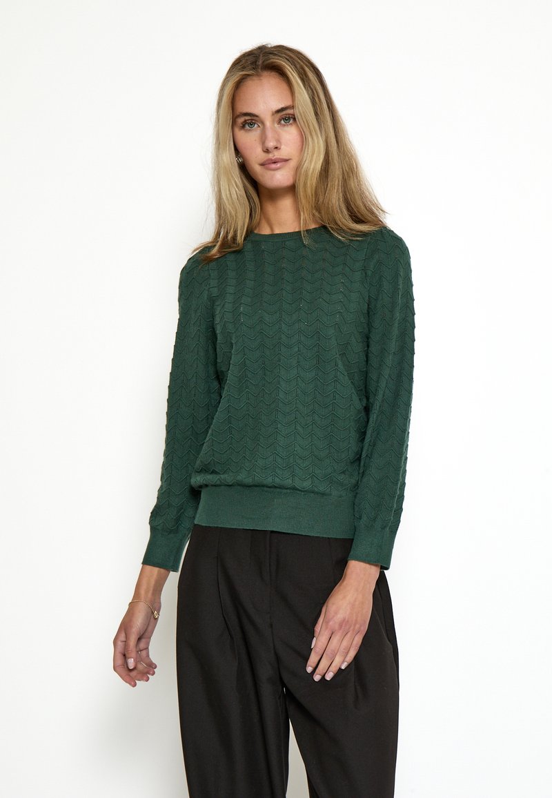 Jersey verde de punto con un patrón de zigzag, puños y dobladillo acanalados, cuello redondo y mangas largas. Combinado con pantalones oscuros.