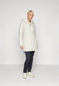 Persona con capelli biondi corti che indossa un lungo piumino bianco, pantaloni scuri aderenti e sneakers bianche, in piedi contro uno sfondo chiaro e uniforme.