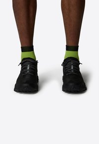Zapatillas deportivas negras con parte superior texturizada, calcetines tobilleros verdes en contraste, suelas de goma y detalles en negro en los cordones y acentos.