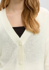 Ljus krämfärgad cardigan i texturerat stickat material med V-ringning och två knappar fram. Tyget har ett något genomskinligt utseende.