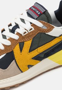 Mehrfarbiger Sneaker aus braunem Wildleder, grünem Mesh, marineblauen Akzenten und einem gelben, strukturierten Überzug. Weiße Schnürsenkel und ein gestreiftes Etikett sind zu sehen.