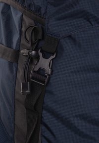 Tela azul marino con una correa negra con hebilla de plástico y un toggle de cordón, mostrando una superficie texturizada con líneas diagonales.