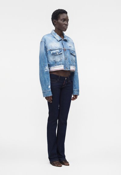 Γυναίκα που φοράει κοντή φθαρμένη μπλε denim μπουφάν και σκούρα μπλε εφαρμοστά τζιν, στέκεται με ουδέτερη έκφραση μπροστά σε απλό φόντο.