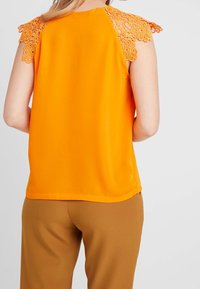 Top arancione senza maniche con accenti in pizzo floreale sulle spalle, texture liscia e vestibilità ampia. Abbinato a pantaloni marroni, vista di schiena.