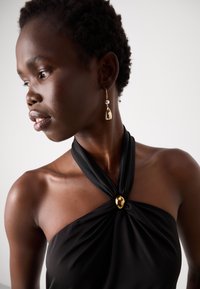 Black sleeveless top with twisted fabric design at the neckline featuring a gold accent. The model wears a gold teardrop earring.