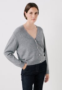 Cardigan grigio in lana con scollatura a V profonda, maniche lunghe e cinque bottoni argentati. Indossato con jeans in denim scuro, texture liscia.