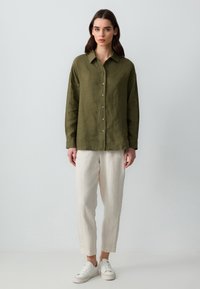 Camicia button-up in lino verde oliva con colletto, abbinata a pantaloni in lino beige chiaro e sneakers bianche. Texture liscia; vestibilità rilassata.