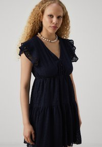 Μπλε ναυτικού φόρεμα φτιαγμένο από υφασμένο ύφασμα με σχέδια οπών, με κοντά ρουστίκ μανίκια και φούστα με στρώσεις και λεπτομέρειες από κουμπιά.