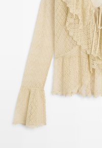 Blusa in pizzo crema a maniche lunghe con polsini svasati, pettorina arricciata e chiusura a fiocco, con un delicato motivo a diamante su tutta la superficie.