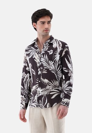 Giovane uomo che indossa una camicia nera con bottoni con stampa bianca di foglie e fiori tropicali e pantaloni beige chiaro, in piedi davanti a uno sfondo semplice.