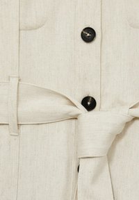 Návrh s off-white button-up odevom z textúrovanej látky, s čiernymi gombíkmi, prednými vreckami a zodpovedajúcim opaskom na páse.