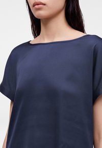 Femme portant un chemisier en satin bleu foncé à manches courtes avec un large col rond sur un fond uni.