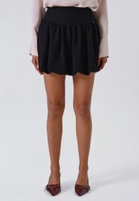 Donna che indossa una mini gonna nera arricciata, una camicia a maniche lunghe beige e tacchi a punta rosso scuro, in piedi contro uno sfondo semplice.