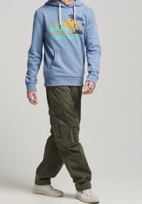 Persona vestida con una sudadera ligera azul con estampado de atardecer tropical y pantalones cargo verde oliva caminando, con zapatillas blancas visibles.