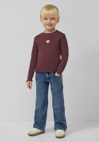 Bordeauxrotes Langarmshirt mit einer glitzernden Herzapplikation, kombiniert mit hellblauen Jeans und weißen Turnschuhen. Lässiger Schnitt mit fransigen Bündchen.