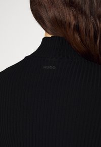 Vrouw met lang bruin haar, gekleed in een zwart geribbelde top met hoge hals en een klein "HUGO" logo op de achterkant van de hals.