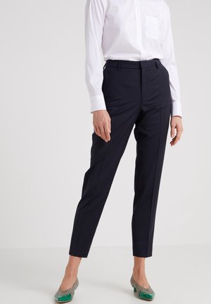 Woman wearing tailored black ankle-length pants, white button-up shirt, and green low-heeled shoes, standing against a plain background.