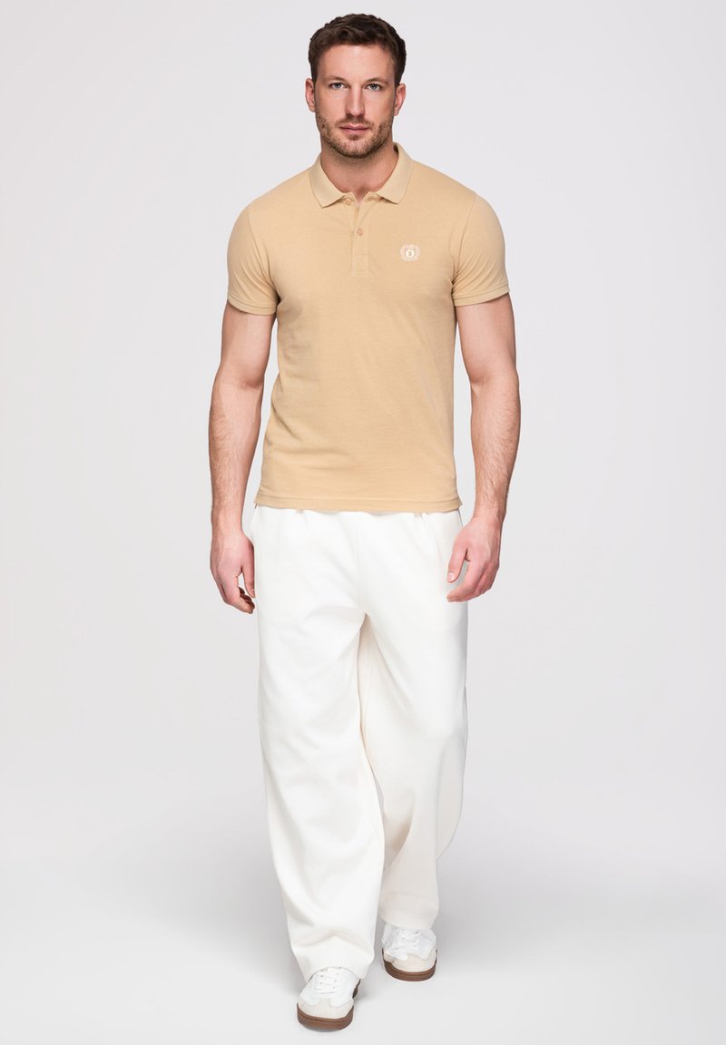 Homme portant un polo beige à manches courtes, un pantalon blanc large et des baskets blanches, debout devant un fond clair uni.