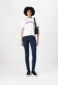 Balta trumpa marškinėliai su "GUESS JEANS" raudonai ruda spalva, derinami su tamsiai mėlynomis siaurėjančiomis kelnėmis ir juodais sportbačiais su baltais padais.