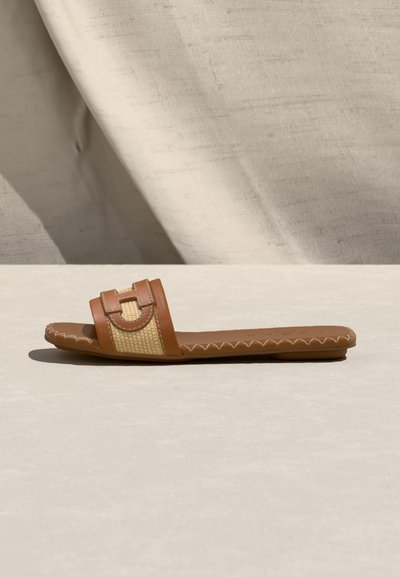 Φλατ καφέ σανδάλι με πλεκτό μπεζ λουράκι και ραμμένη σόλα, παρουσιάζεται μπροστά σε φωτεινό υφασμάτινο φόντο πάνω σε ουδέτερη επιφάνεια.