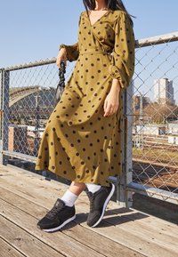 Femme en robe à pois vert olive portant des baskets noires et des chaussettes blanches, debout sur une plateforme en bois près d'une clôture en grillage dans un environnement urbain.