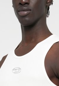 Άσπρο αμάνικο τοπ με στρογγυλή λαιμόκοψη, ραφήρτη υφή και μικρό ασημί λογότυπο στο στήθος. Ορατή λεπτομέρεια από απαλό ύφασμα.