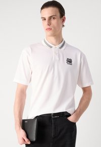 Witte polo met een grijze gestreepte kraag, voorzien van een zwart logo op de linkerborst. Model houdt een zwarte clutch vast en draagt een donkere spijkerbroek.