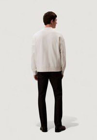 Bílá mikina s žebrovanými manžetami a lemem, kombinovaná s tmavými džínami. Model je zobrazen zezadu na jednoduchém světlém pozadí.