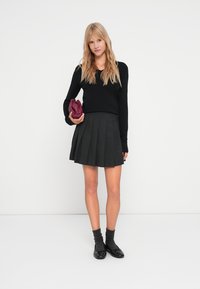 Svart långärmad tröja kombinerad med en svart plisserad kjol. Modellen håller en vinröd clutch och har på sig mörka strumpor med svarta skor.